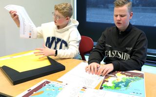 Twee jongens, één in een witte hoodie, de ander in een zwart vest, zitten aan een tafel. Voor zich hebben ze een kaart van Nederland.