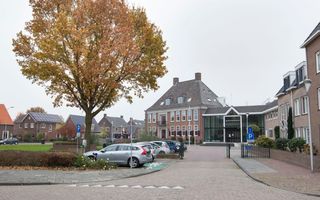 Groot gebouw met parkeerplekken, auto’s en een boom ervoor. 