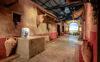 Foto van een straatje met zandkleurige panden links en rechts. Langs de muren staan grote kruiken. Aan het eind is een poort met uitzicht op de landelijke omgeving.