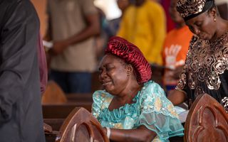 Een donkere vrouw zit in een kerkbank. Ze heeft haar ogen gesloten en heeft een verdrietige uitdrukking op haar gezicht. 