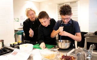 Twee jongens zijn eten aan het koken. Een vrouw geeft instructies. Op de voorgrond schalen met eten. 