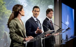 Vrouw met groen jasje en twee mannen in pak staan achter spreekgestoeltes. 