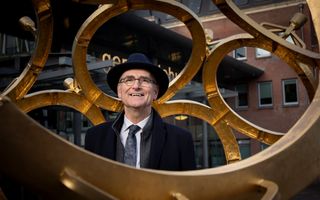 Een oudere man met donkerblauwe hoed en een jas in dezelfde kleur staat in een kunstwerk met gouden ringen. In de achtergrond een gebouw. Op de gevel zijn nog een paar letters te zien van het woord ‘gemeentehuis’.