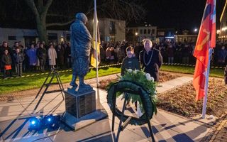 Man met ambtsketen en jongen kijken naar krans naast beeld.
