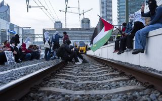 Mensen met doeken om het hoofd zitten op de rails, met Palestijnse vlag. In de verte is een trein zichtbaar.