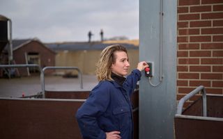 Een blonde vrouw in een blauwe overall duwt op een rode knop aan een muur. Op de achtergrond zijn stallen te zien.