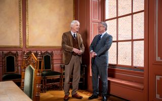 Twee grijzende mannen in pak staan in een ruimte met donkerroze lambrisering. Achter hen staan enkele klassieke eikenhouten stoelen met fluwelen bekleding.