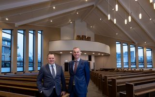 Twee mannen in pak, lachend, in het gloednieuwe kerkgebouw. Voorin de kansel, aan weerszijden hoge ramen.