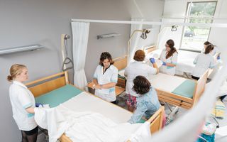 Studenten en docenten in wit uniform staan rond ziekenhuisbedden waarop ze lakens leggen.