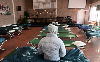 Man met jas en capuchon, op de rug gezien. Hij zit op een veldbed in een kerkzaal vol veldbedden.