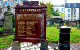 Donkerrood monument met goudkleurige namen en jaartallen in een parkje tussen historische panden.