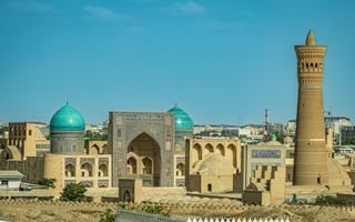 Een oud, bruin gebouwencomplex met turqoise koepels en een zandbruine minaret, tegen een felblauwe lucht. 