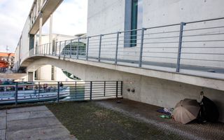 Een dakloze zit weggedoken achter een paraplu onder een een grijze betonnen brug. 