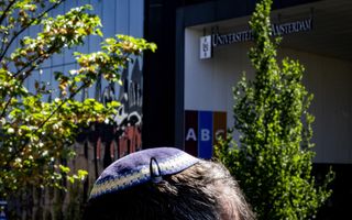 Een blauw-paarskleurig keppeltje dat met een haarspeltje vast zit op het hoofd van een verder onzichtbare man. Achter hem een gebouw van de Universiteit van Amsterdam.