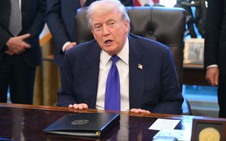 Een man in blauw kostuum en met een blauwe stropdas zit achter een bureau.