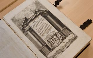 Opengeslagen Bijbel op een houten tafel bij het titelblad met daarop een afbeelding van een gravure.
