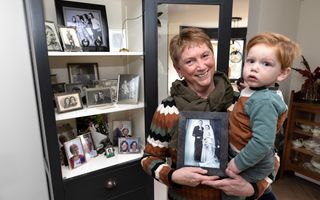 Een vrouw met een jongetje op de arm laat een oude trouwfoto zien. Naast haar een vitrinekast met familiefoto’s.