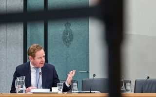 Man in pak zit in een commissiezaal van de Tweede Kamer en gebaart met zijn hand.