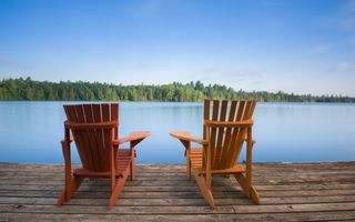 Twee houten stoelen die uitkijken op een meer. In de verte is een bomenrij te zien.