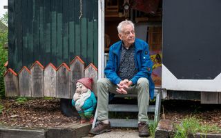 Een man met grijs haar zit op een trapje in de deuropening van een soort schaftkeet en kijkt schuin opzij. Naast hem staat een kabouterfiguur.