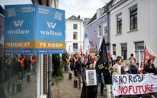 Op de voorgrond is een blauw bord te zien waarop staat: te koop. Op de achtergrond lopen studenten met een spandoek: no roof, no future – geen dak, geen toekomst. 