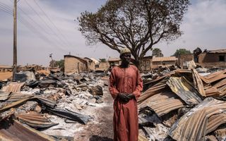 Een man in een lang, rood gewaad staat tussen de verbranden resten van golfplaten woningen.