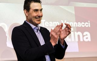 Een Italiaanse man met donker haar en donker pak met lichtblauw overhemd klapt lachend in zijn handen. 