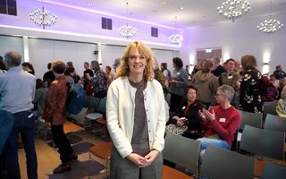 Een vrouw met halflang, blond haar en een wit vest staat voor een zaal vol mensen, die zitten of staan.  