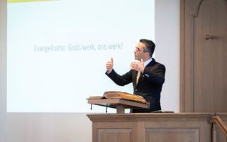 Een man in een zwart pak spreekt met armgebaren op een preekstoel, boven een open kanselbijbel. Op de achtergrond staat tekst op een beamerscherm.