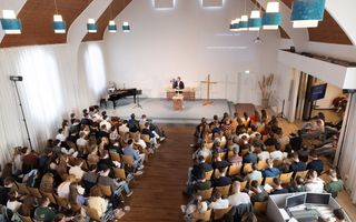 Een kerkzaal vol mensen die op stoelen zitten, van bovenaf gezien. Voorin staat een man achter een katheder op een podium.