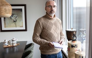 Man met bril, bruine coltrui en spijkerbroek staat bij een raam met een boekje in zijn handen. In het raamkozijn staan een djembé en wat exotische voorwerpen.