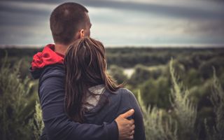 Een man en een vrouw kijken uit over een natuurgebied. Ze hebben de armen om elkaar heen geslagen.