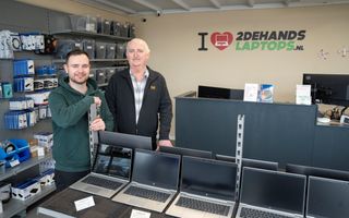 Twee mannen staan in een showroom van tweedehands laptops tussen beeldschermen en laptops, met op de achtergrond computeronderdelen.