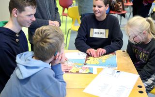 Vier kinderen en jongeren zitten om een tafel waar topografische kaarten van Nederland liggen.