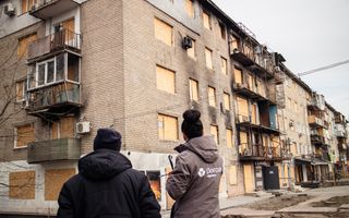 Man en vrouw met jacks, van achteren gezien, bij gebouw waar ramen dichtgespijkerd zijn.