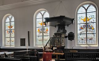 Het interieur van een historische kerk. De preekstoel staat tussen fraai gekleurde boogramen.
