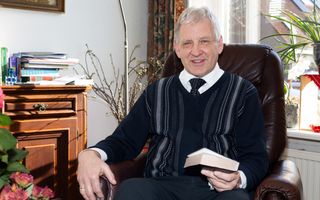 Predikant in zwarte trui met stropdas zit in bruine stoel met Bijbel in zijn hand.