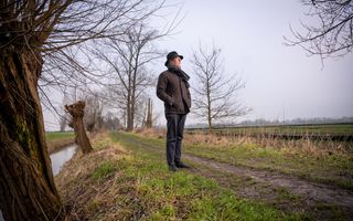 Een man op leeftijd in winterkleding, met een zwarte hoed op het hoofd, staat op een boerenweggetje tussen de weilanden en kijkt naar de lucht. In de berm staan knotwilgen langs een sloot.