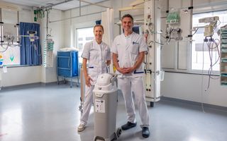 Een man en een vrouw in witte artsenkleding achter een grijze luieremmer. Op de achtergrond snoeren en apparatuur. 