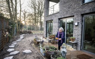 Oudere vrouw met paarse sjaal zet iets neer op het terras van haar appartement. 