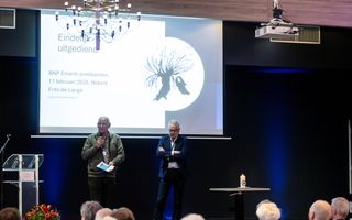 Twee mannen staan op het podium, achter hen een scherm met daarop de woorden ”Eindelijk uitgediend”.