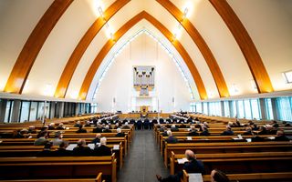 Interieur van een modern kerkgebouw met daarin bezoekers in banken en voorin een kring van tafels met veertig mannen daaromheen.