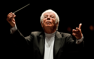 Een oudere dirigent aan het werk met een dirigeerstokje in zijn hand. Hij heeft grijs haar, een bril, draagt een witte koltrui met daaroverheen een zwart jasje.