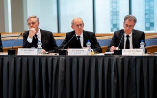 Drie mannen in zwarte pakken, met bril, zitten achter microfoons aan vergadertafels.