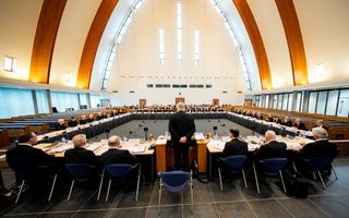 Vergadering van 41 personen rond tafels in een modern kerkgebouw; de zeven mensen van het vergaderbestuur worden hier op de rug gezien.