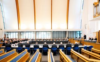 41 mannen in zwarte pakken zitten in een modern kerkgebouw met lichte kleuren rond vergadertafels.