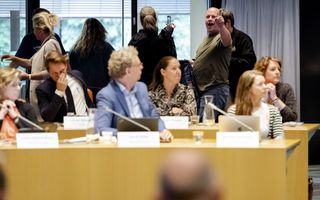 Een kale man steekt dreigend zijn vinger op en schreeuwt iets door de raadszaal. Op de voorgrond zitten de gemeenteraadsleden, waarvan een de hand voor zijn mond houdt. 