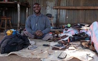 Een donkergetinte man zonder benen zit glimlachend op de grond tussen zijn schoenmakersgereedschap in een werkplaats gebouwd van golfplaten en hout.