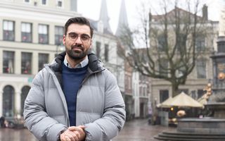 Man met donker kapsel en grijze jas in het centrum van een oude stad.