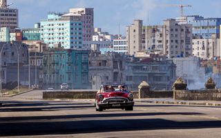 Een rode oldtimer rijdt over een vrijwel lege weg. In de verte zijn gebouwen in slechte staat zichtbaar.